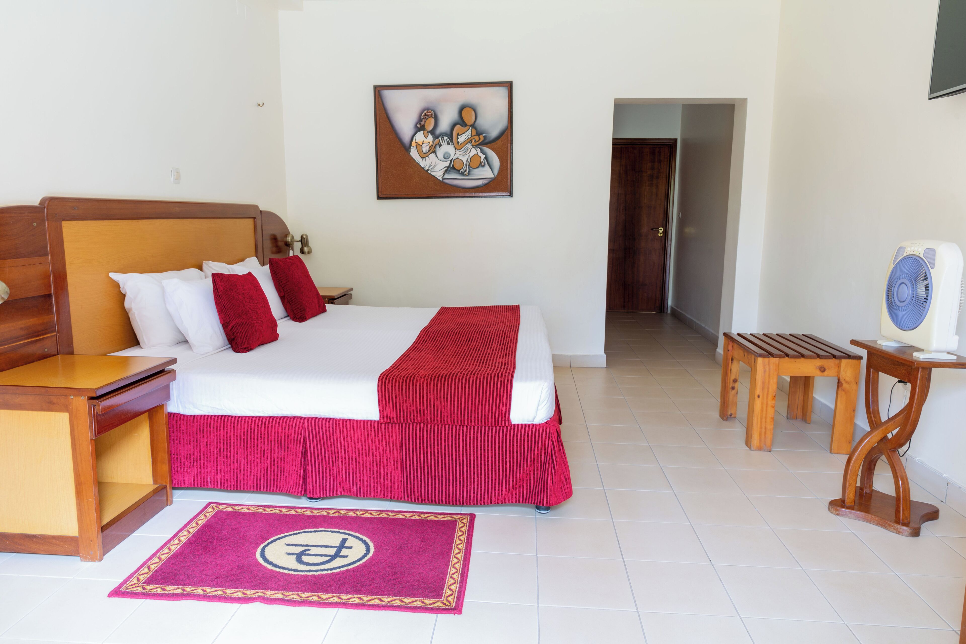 Habitación de hotel con una cama, mesitas de noche de madera, colcha roja, un cuadro en la pared, un banco de madera y un ventilador.
