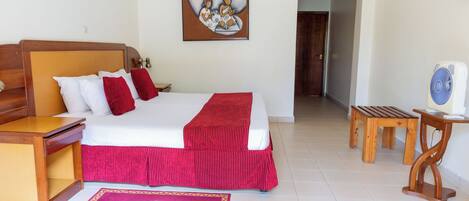 Habitación de hotel con una cama, mesitas de noche de madera, colcha roja, un cuadro en la pared, un banco de madera y un ventilador.