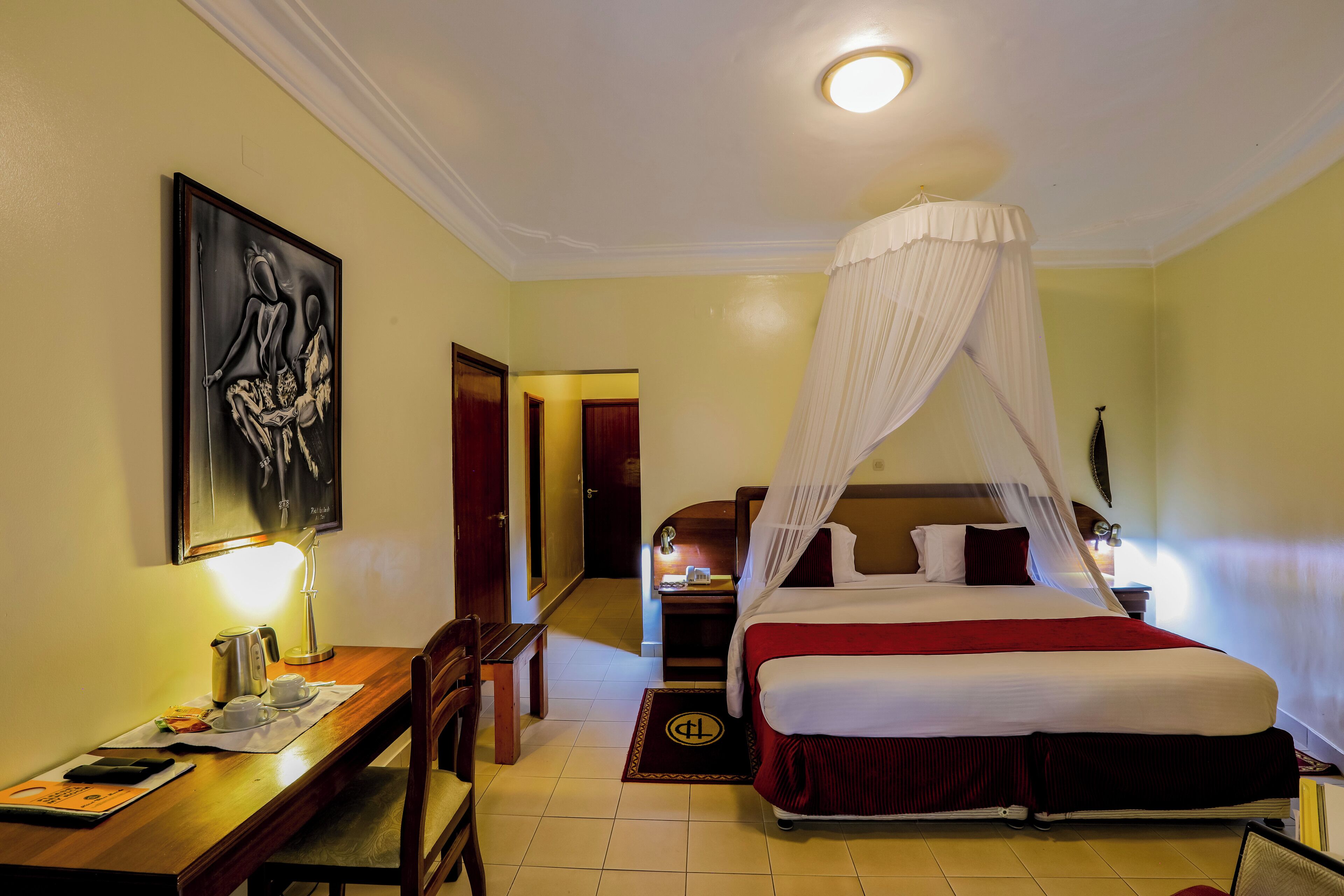 A hotel room with a bed, a desk with a lamp, a chair, and a painting on the wall.