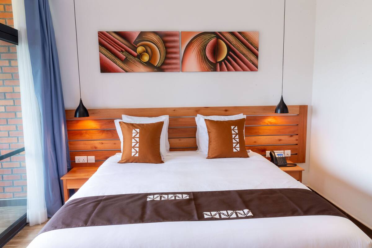 A hotel room with a wooden headboard, two brown pillows, a white bedspread, and a brown blanket. There are two wall-mounted artworks above the bed.