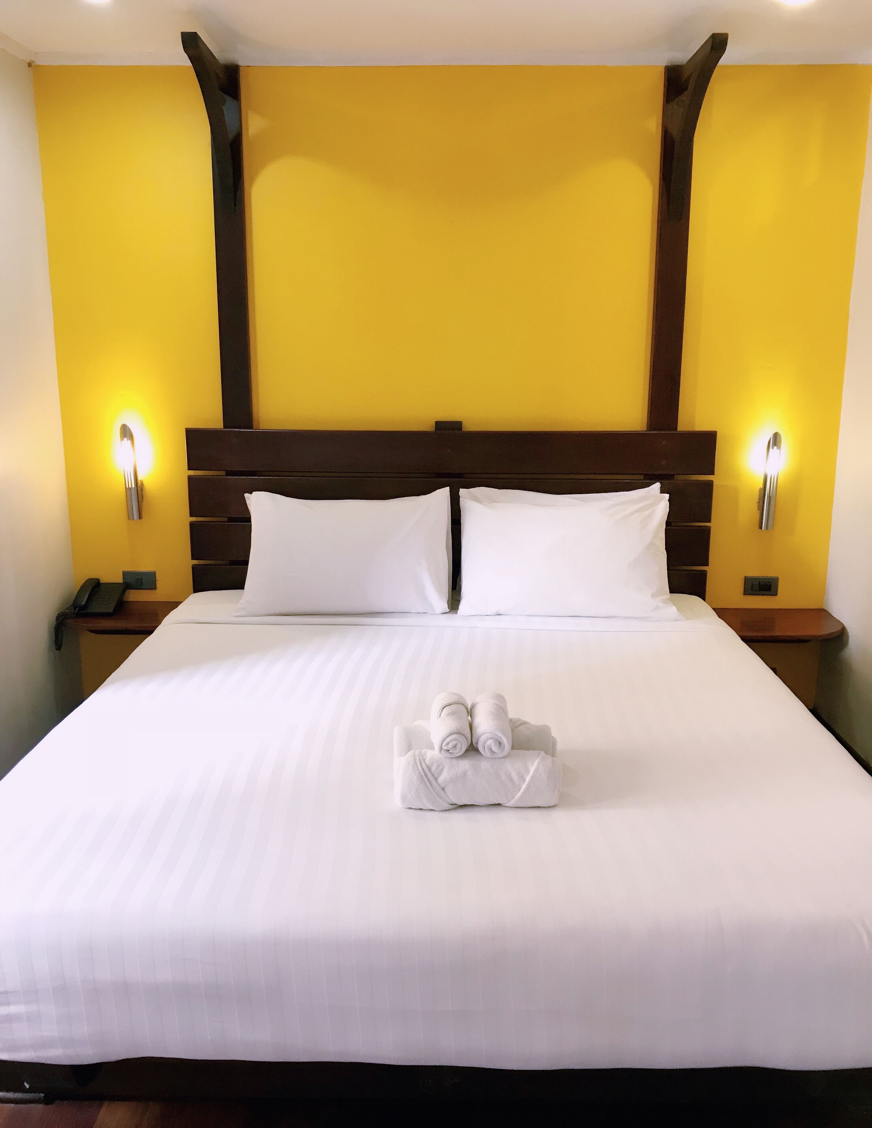 A large bed with white linens, two pillows, and a towel arrangement. There are two bedside tables with lamps and a phone on one. The room has yellow walls and a wooden headboard.