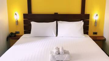 A large bed with white linens, two pillows, and a towel arrangement. There are two bedside tables with lamps and a phone on one. The room has yellow walls and a wooden headboard.