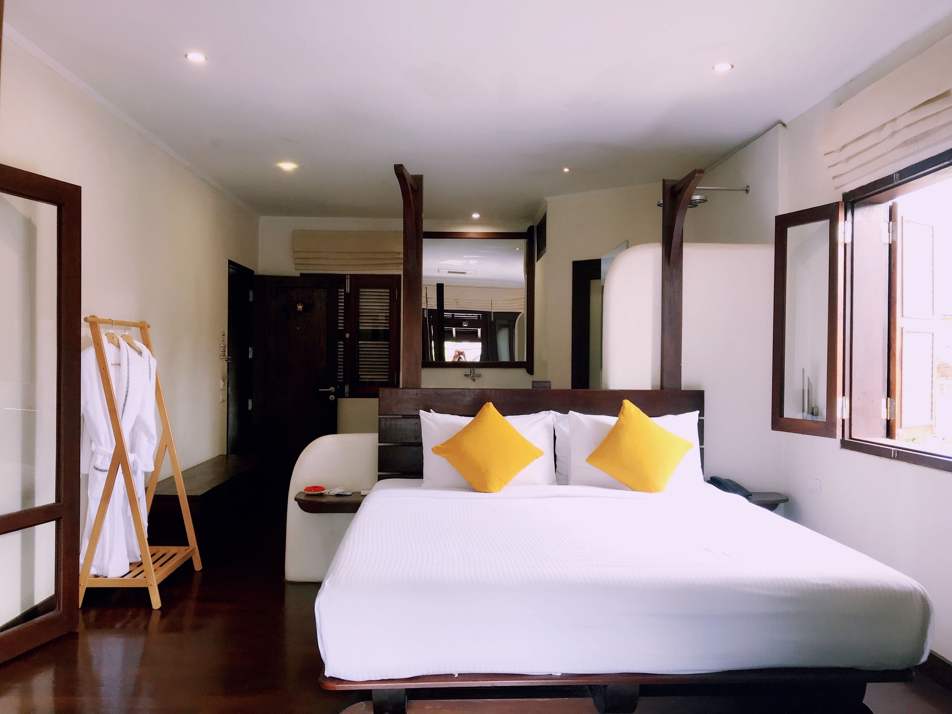 Ein Hotelzimmer mit einem großen Bett, einem Holzkopfstück und zwei gelben Kissen. Ein Holzgestell mit Bademänteln und ein Spiegel an der Wand sind ebenfalls vorhanden.