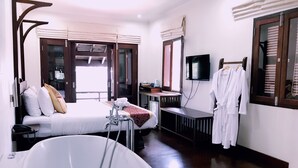 A hotel room with a large bathtub, a bed with pillows, a desk with a TV, and a robe hanging on the wall. - Ancient Luang Prabang Hotel (Luang Prabang)