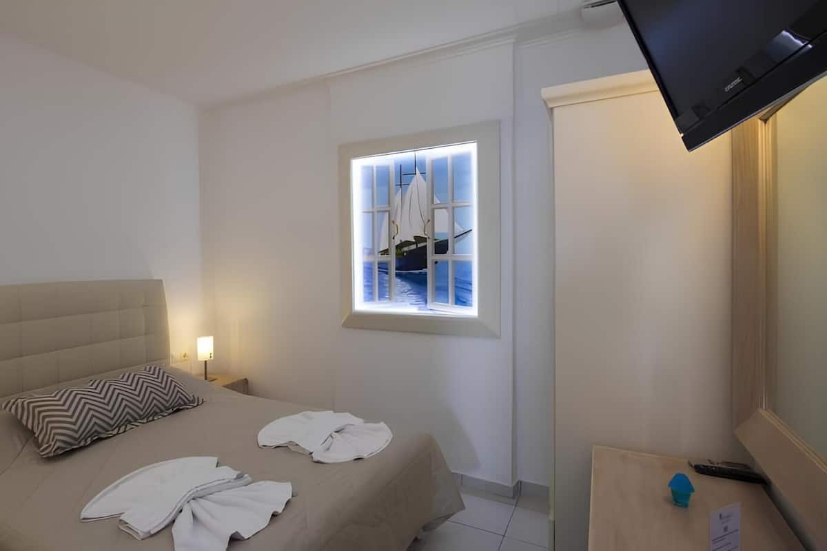 Habitación de hotel con cama, mesita de noche, televisión, ventana y vista a un velero, y una pequeña mesa con control remoto.