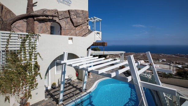 Zona piscina con vista sul mare, un edificio bianco con facciata a roccia e una struttura a pergola.