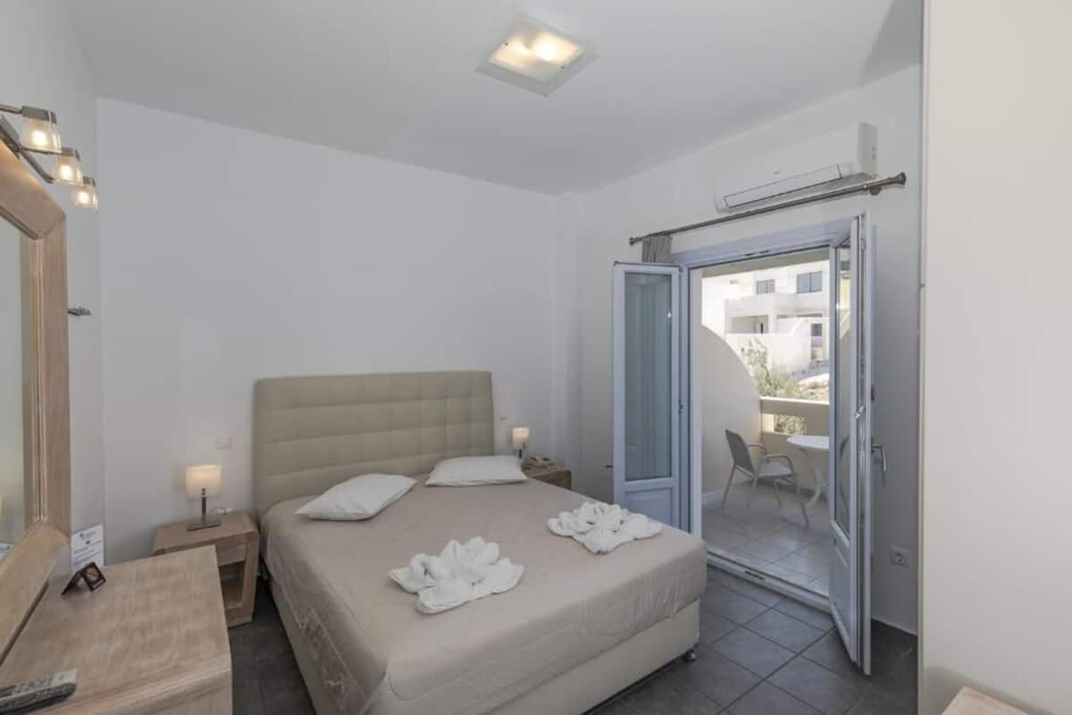 Habitación de hotel con cama, mesitas de noche, espejo y puerta abierta a un balcón con una silla.