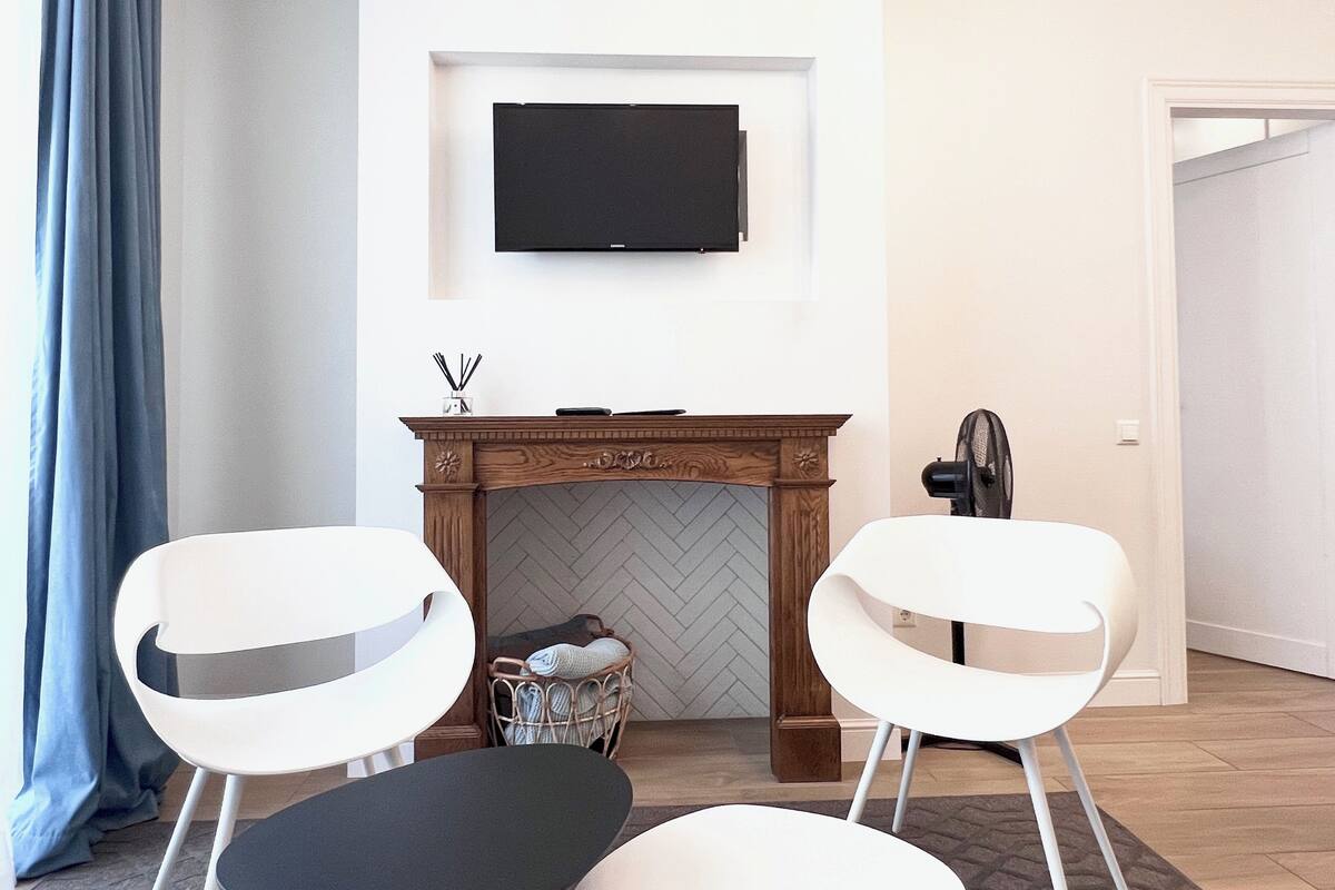A modern living room with a fireplace, two white chairs, a TV mounted above the fireplace, and a fan to the right.