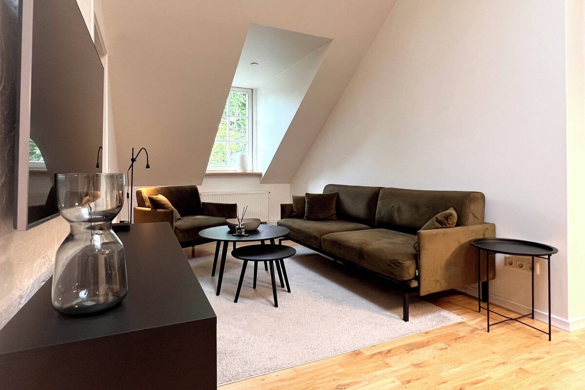 A modern loft apartment with a sloped ceiling, a sofa, two small tables, and a glass sculpture on a dark table.