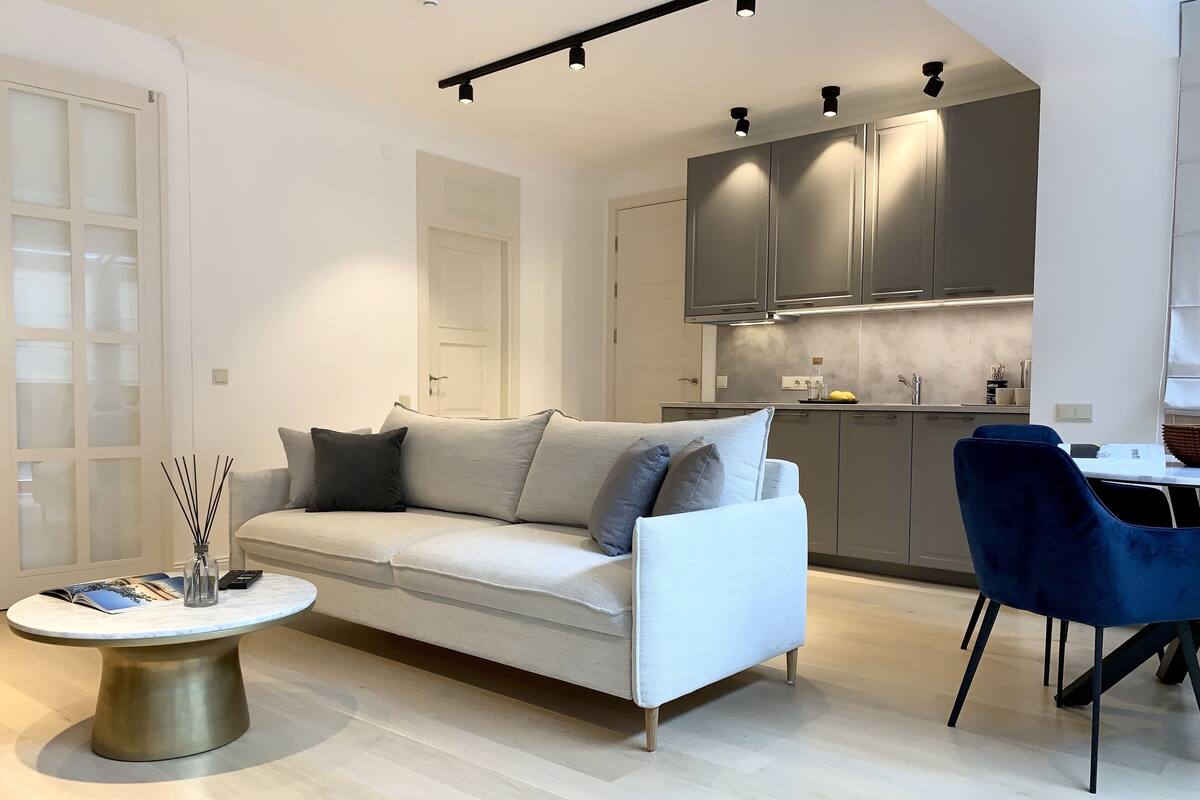 A modern living room with a white sofa, a round coffee table, and a blue chair. A kitchen with grey cabinets and a dining area is visible in the background.