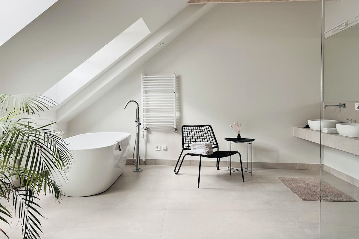 A modern bathroom with a freestanding bathtub, a black chair, a small table with a vase, and a large mirror.