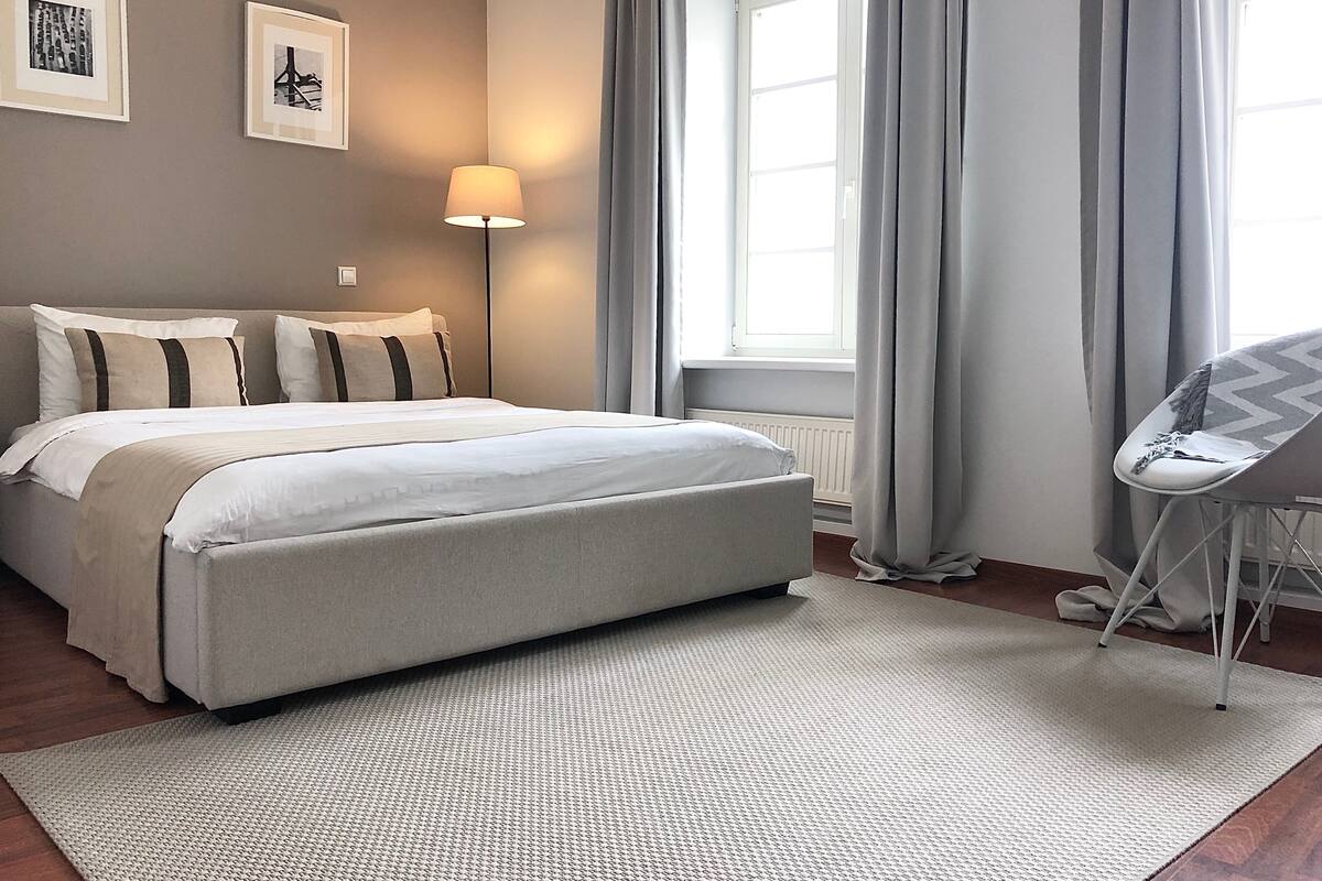 A neatly made bed with a white duvet and two pillows, a standing lamp, a window with curtains, and a chair.