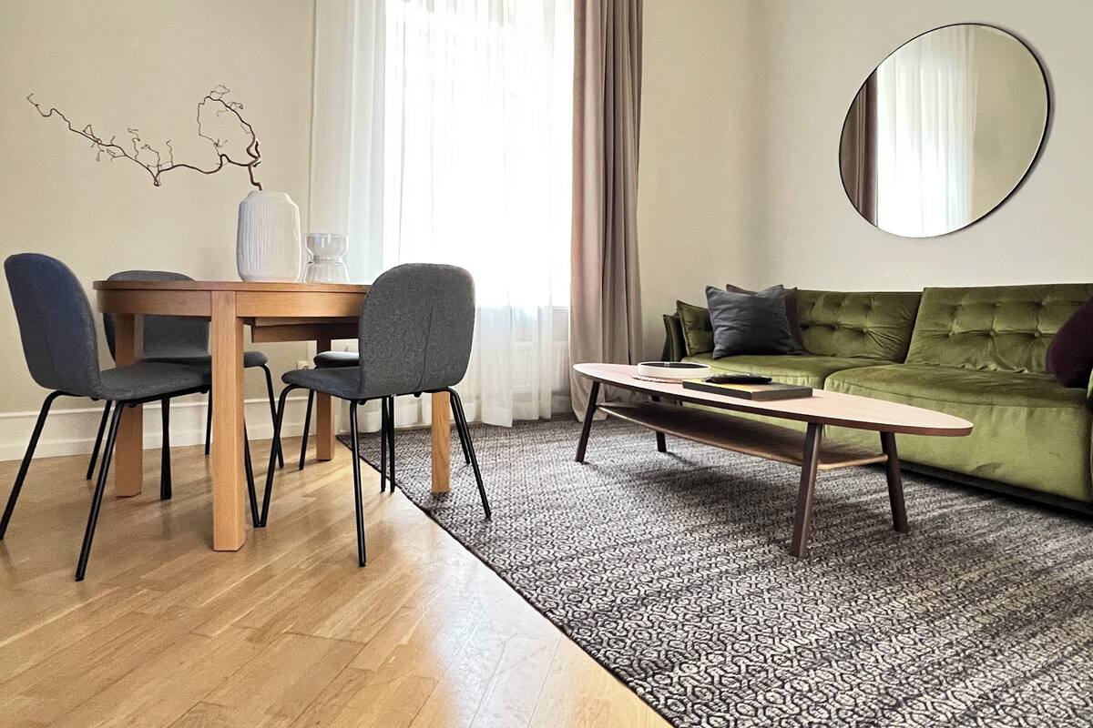 A modern living room with a wooden dining table, two chairs, a green sofa, a coffee table, and a large mirror on the wall.