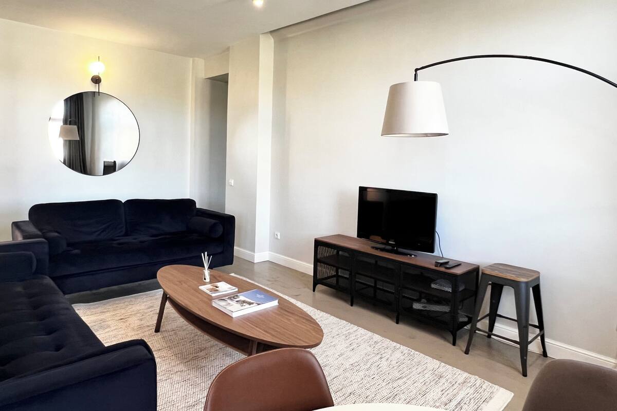 A modern living room with a dark sofa, a wooden coffee table, a TV on a wooden stand, and a round mirror on the wall.