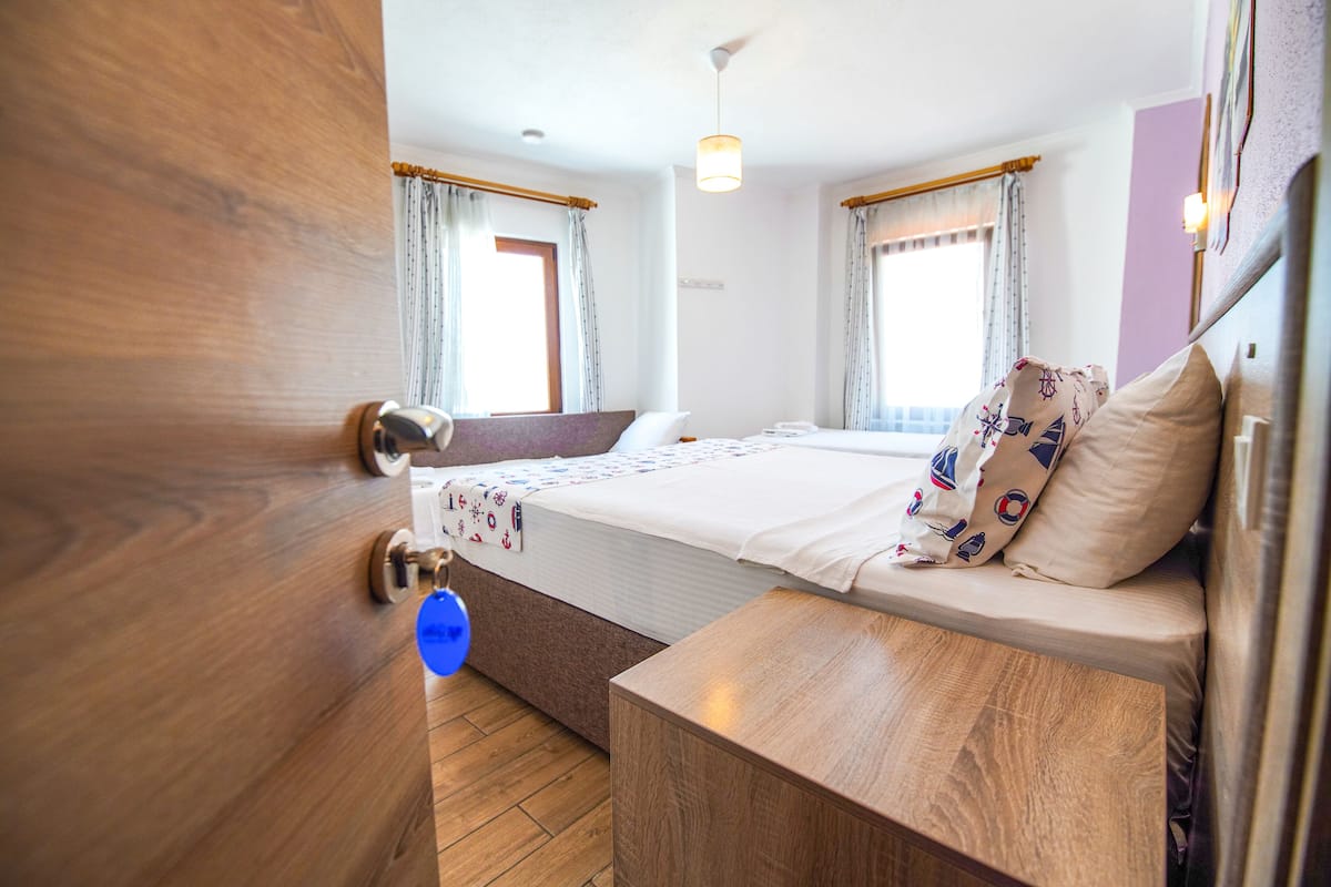 A hotel room with a wooden door, a bed with a patterned bedspread, a bedside table, and a window with curtains.