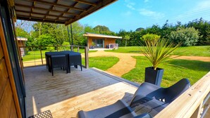 Una terraza de madera con dos sillas reclinables negras y una mesa, con vista a un área verde con un sendero y un edificio pequeño al fondo.
