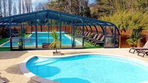 Una piscina con cerramiento de vidrio, sillas de descanso y una sombrilla de paja.