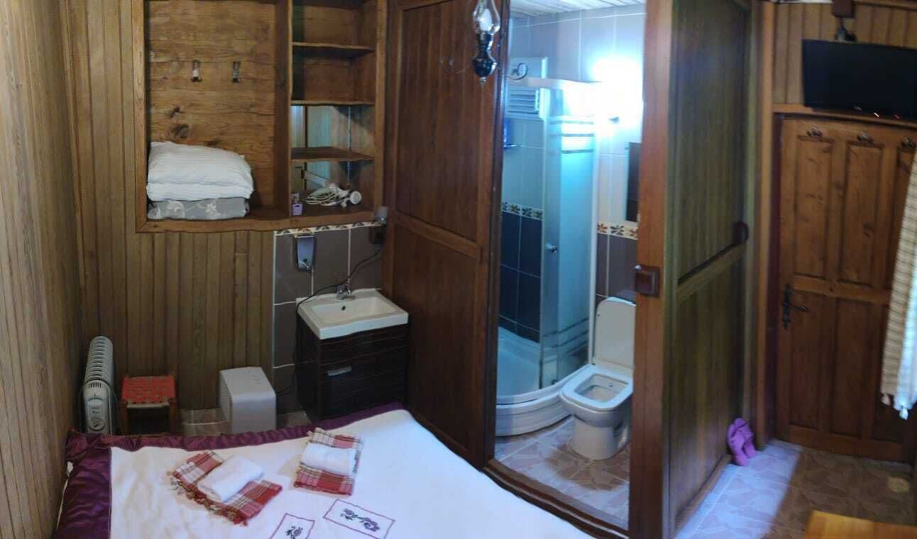 A compact bedroom with a wooden wardrobe, a small bedside table, a bed with a white and red blanket, and a view of a bathroom with a shower and toilet.