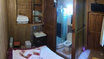 A compact bedroom with a wooden wardrobe, a small bedside table, a bed with a white and red blanket, and a view of a bathroom with a shower and toilet.