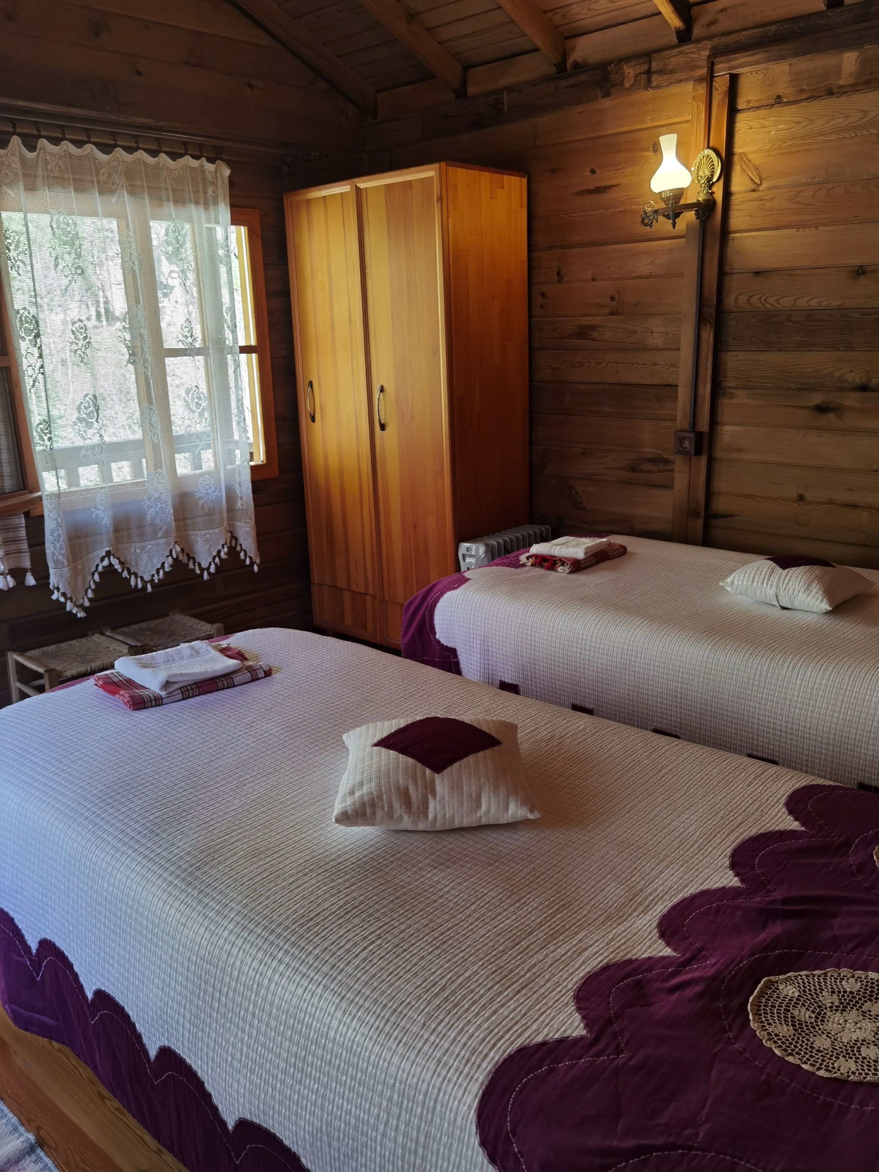 A wooden cabin room with two beds, a wardrobe, and a window with lace curtains.