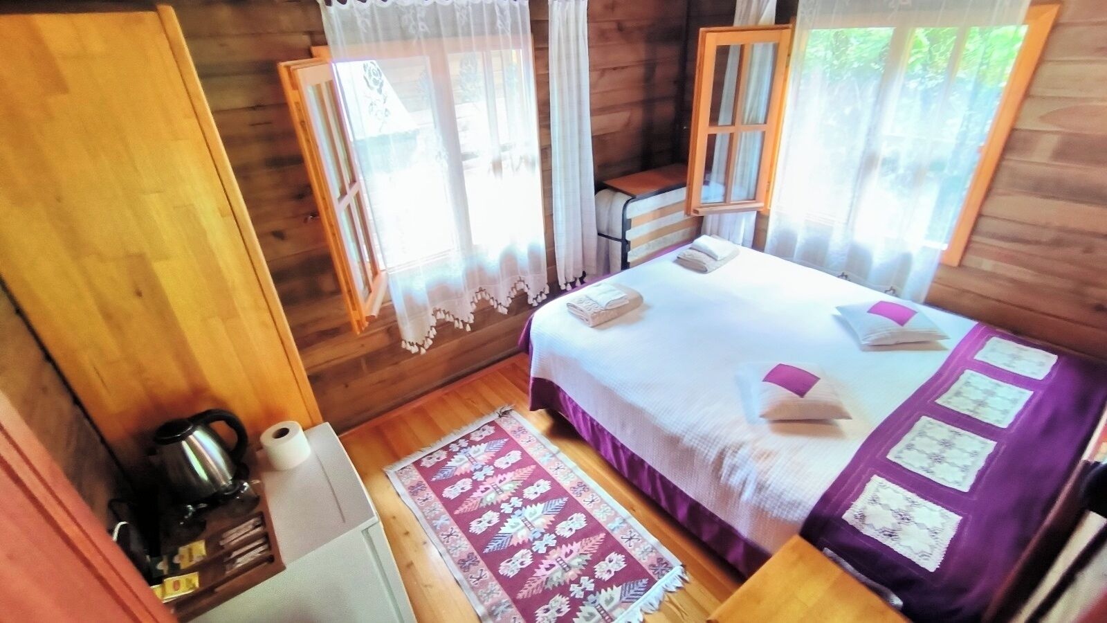 Une chambre dans un chalet en bois, avec un lit, une petite table et une bouilloire, ainsi qu’une fenêtre avec des rideaux.