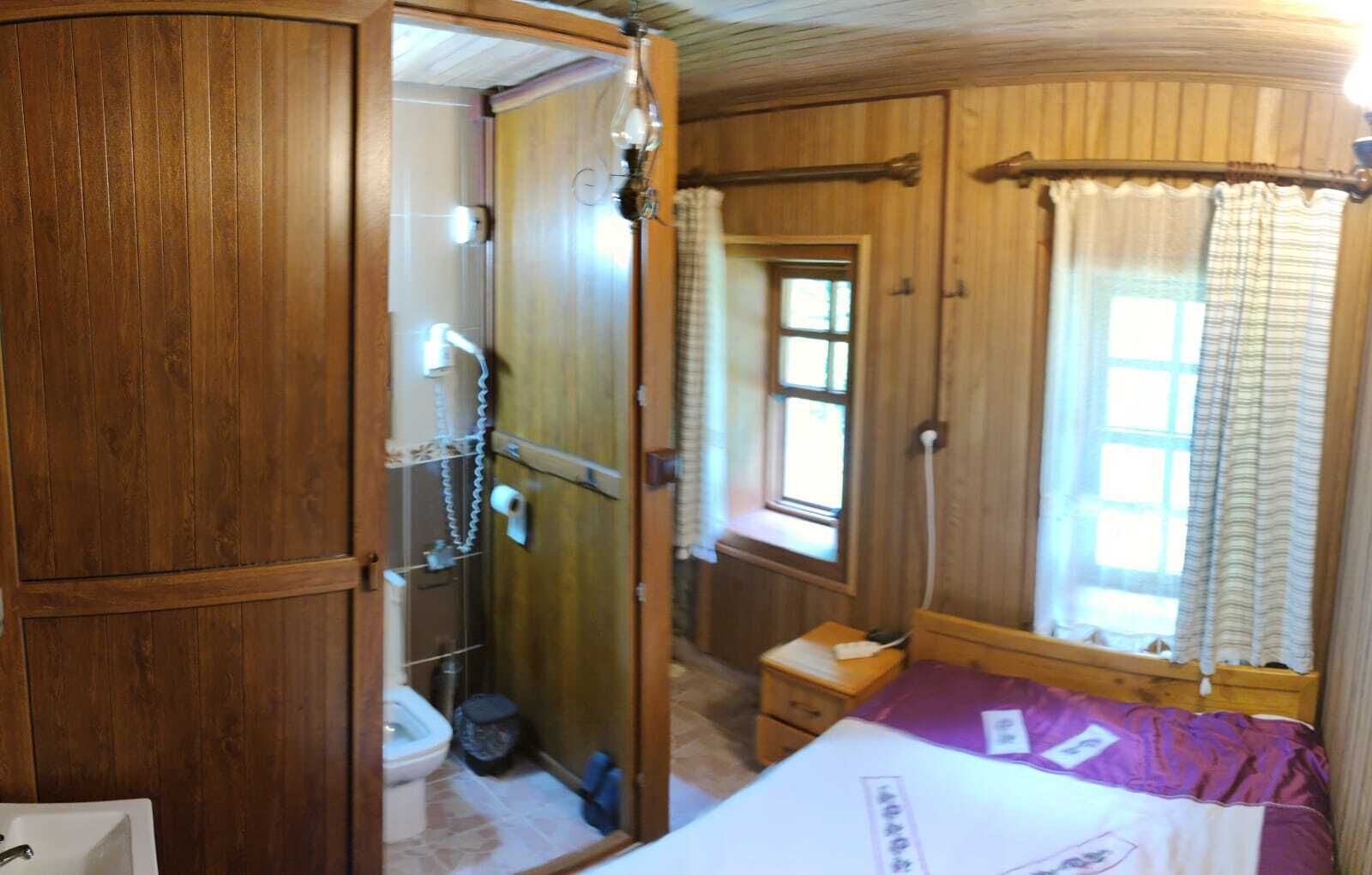 Une pièce de taille modeste avec une porte en bois, une petite fenêtre avec des rideaux, un lit recouvert d’une couverture violette et une armoire en bois.