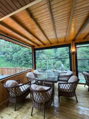 A wooden deck with wicker furniture and large windows overlooking a forest. - Mencuna Konaklari (Arhavi)