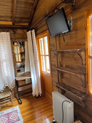 A wooden cabin interior with a television mounted on the wall, a radiator, and a window with curtains. - Mencuna Konaklari (Arhavi)