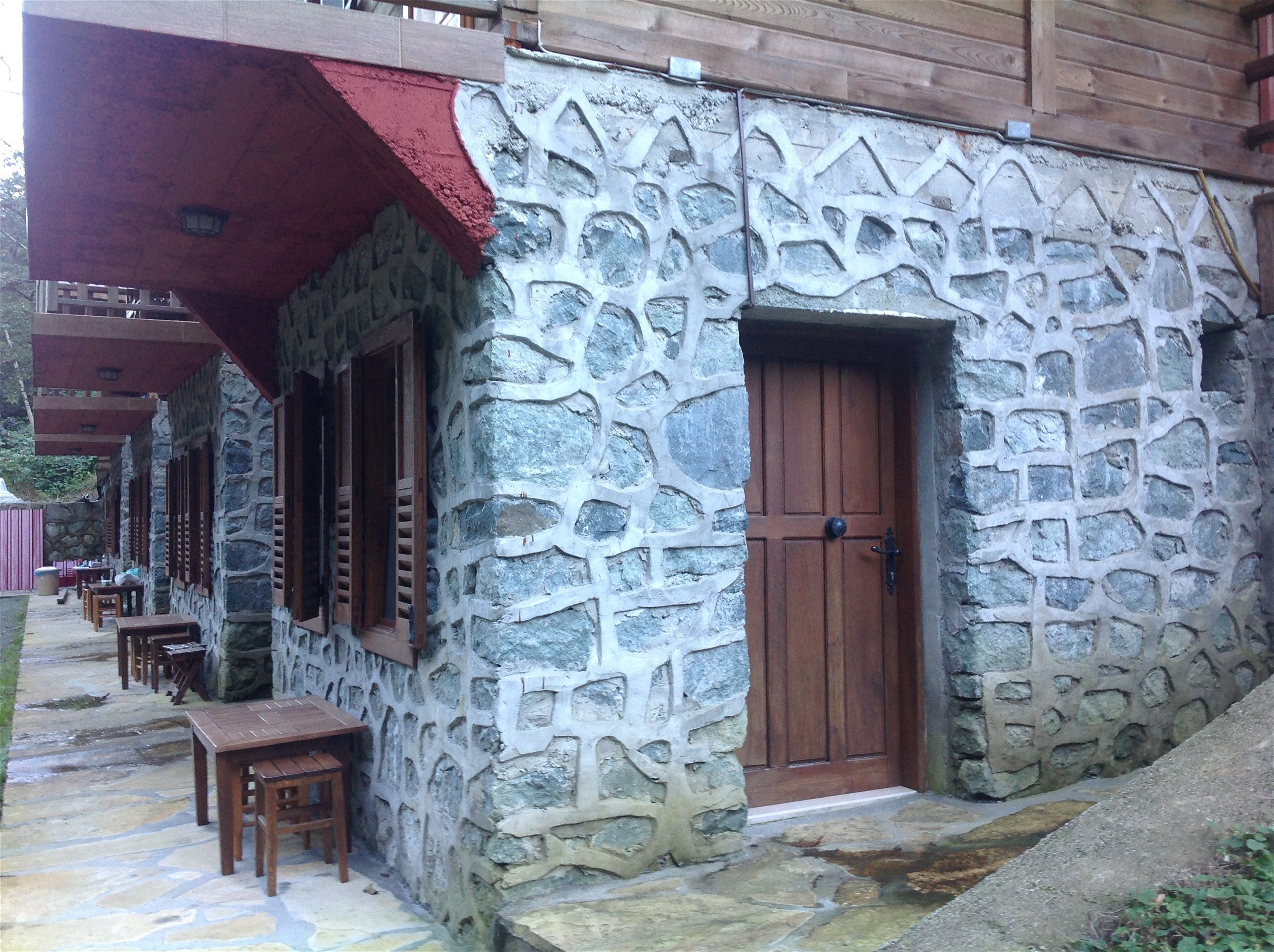 A stone building with a wooden door and windows, outdoor seating with tables and chairs, and a red awning.