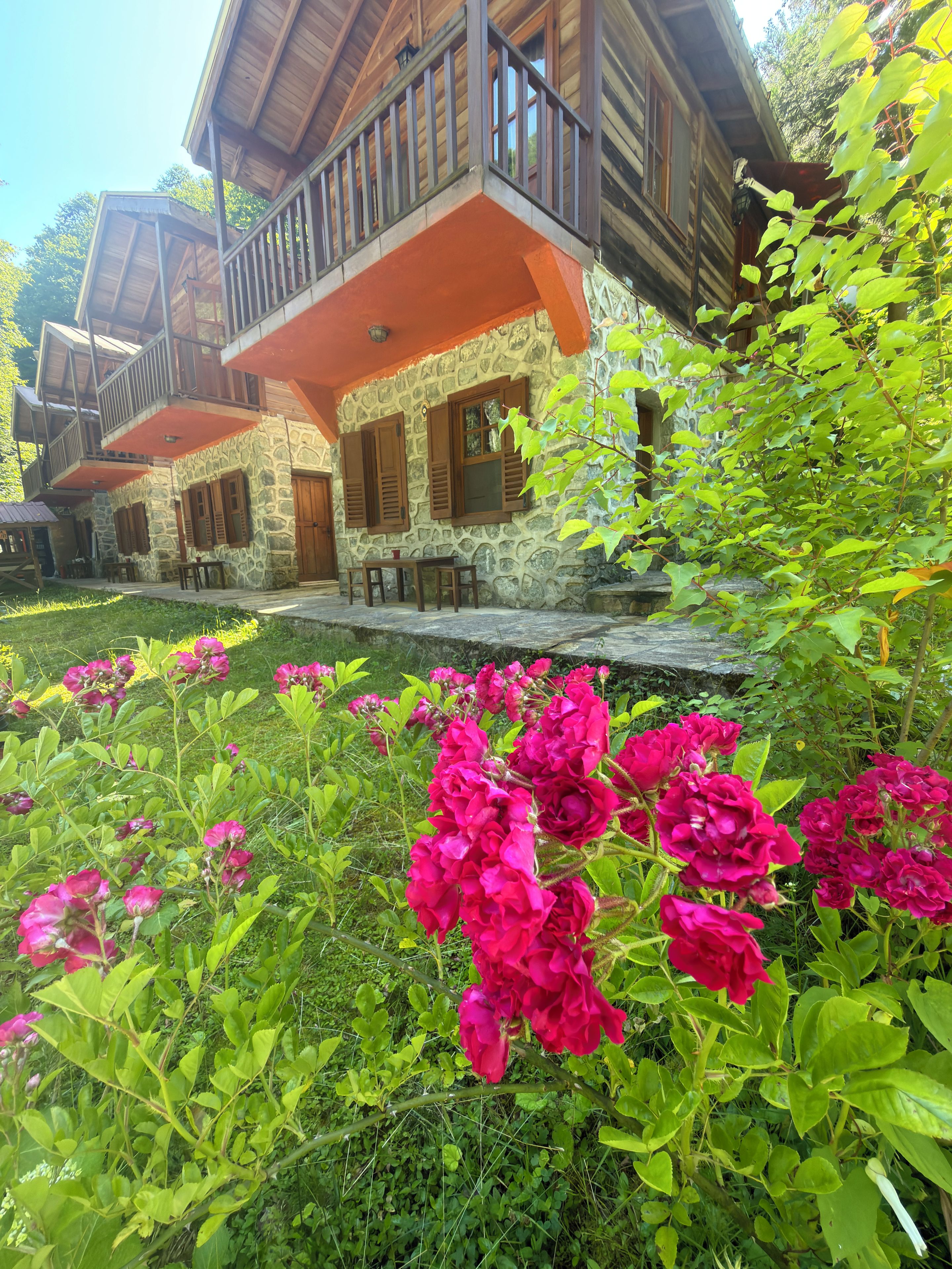 A rustic stone building with a wooden balcony, surrounded by lush greenery and vibrant flowers.