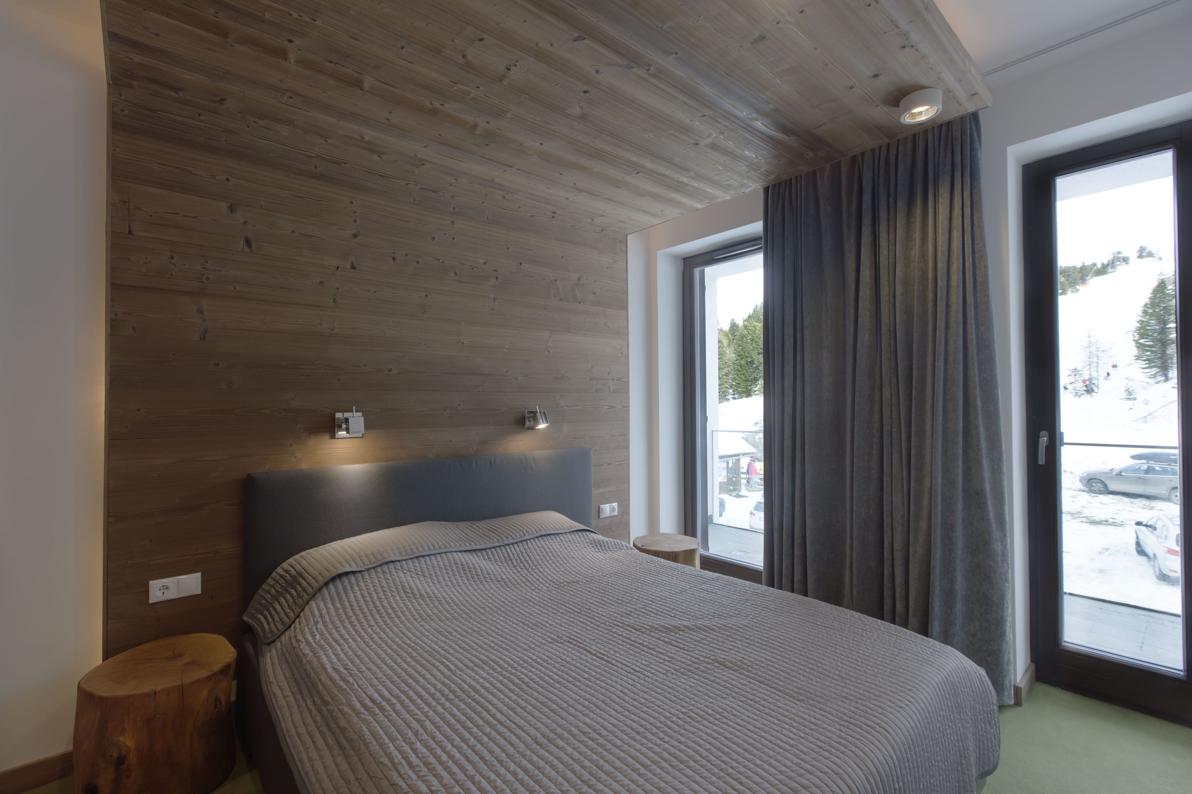 Une chambre à coucher moderne avec un plafond en bois, un grand lit doté d’une tête de lit, une table de chevet et une fenêtre avec des rideaux.