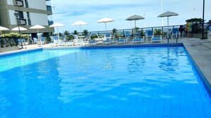 A swimming pool with lounge chairs and umbrellas, surrounded by a fence and a building. - Flat Barrabella (Rio de Janeiro)