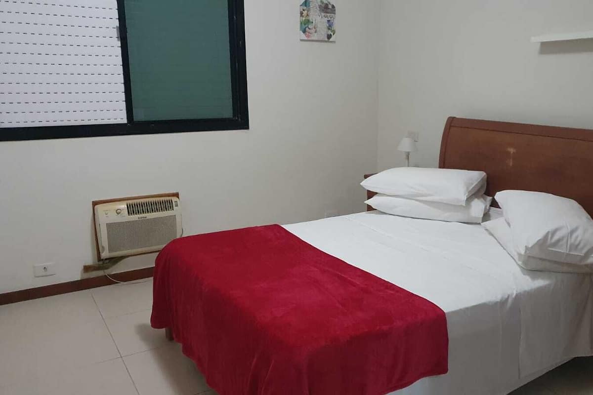 A bedroom with a bed, a red blanket, white pillows, a wooden headboard, a bedside table, a lamp, and a wall-mounted air conditioner.