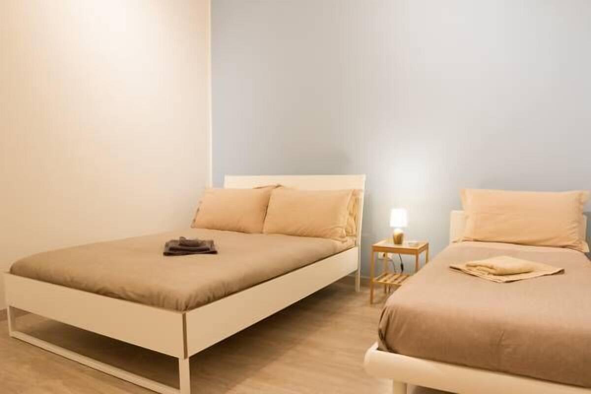 A bedroom with two beds, each with a white frame and beige bedding. There is a small bedside table with a lamp on one side.