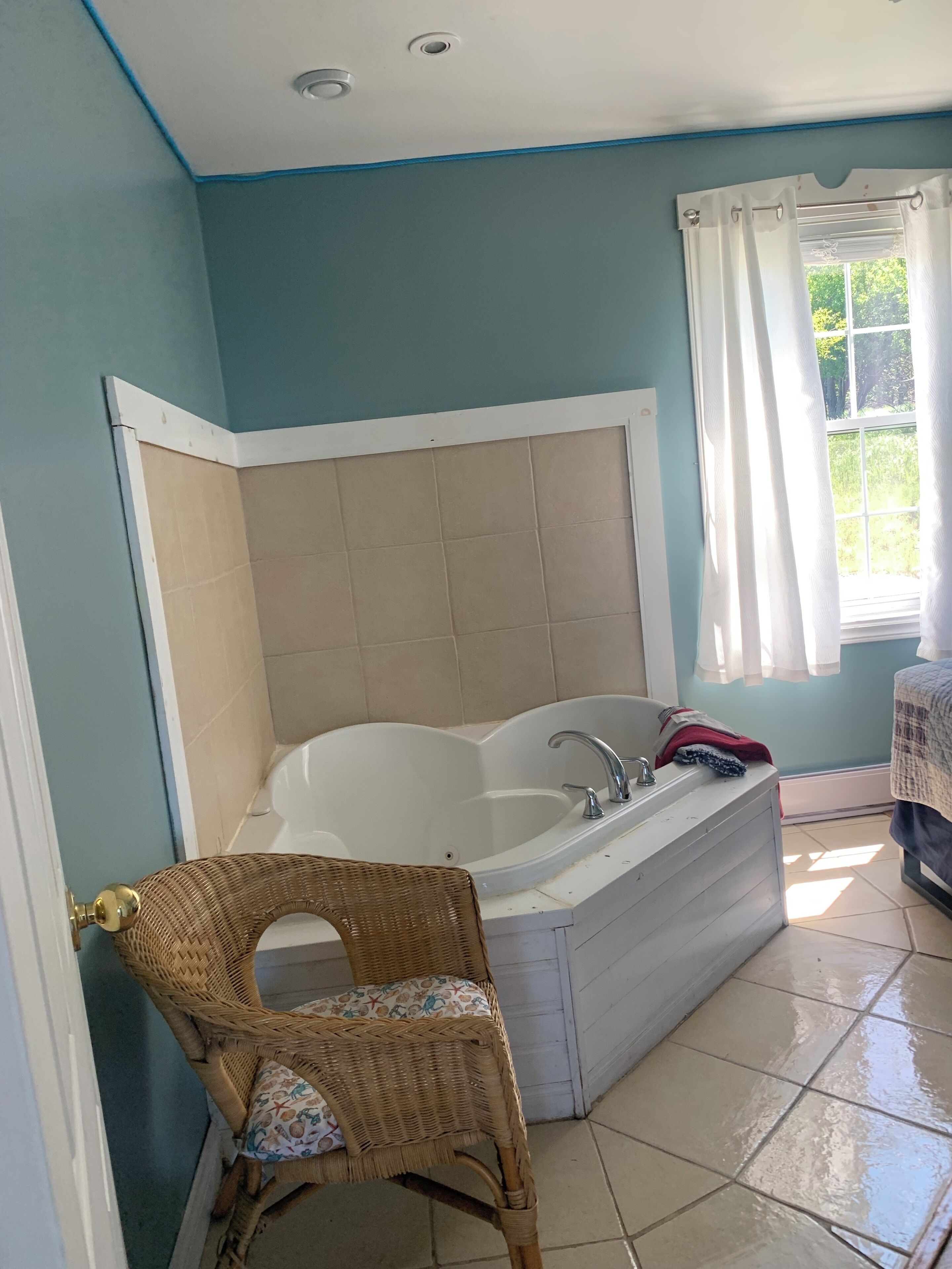 Un baño con bañera, una silla de mimbre y una ventana con cortinas.