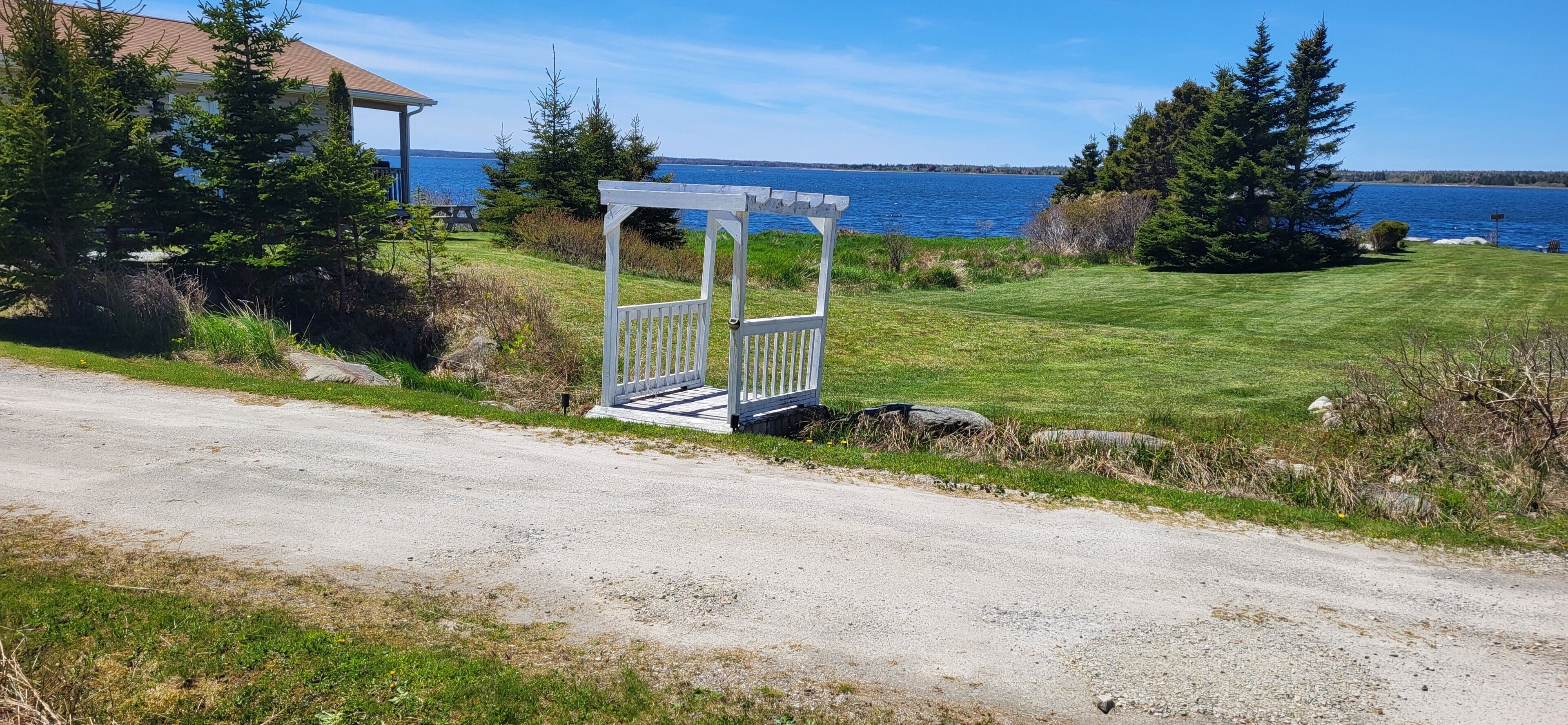 A gravel path leading to a white wooden structure near a body of water, with a building and trees in the background.