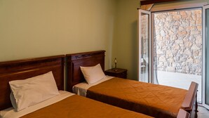A hotel room with two beds, a wooden headboard, a nightstand, and a stone wall visible through the glass door. - Epidavros Seascape (Epidaurus)