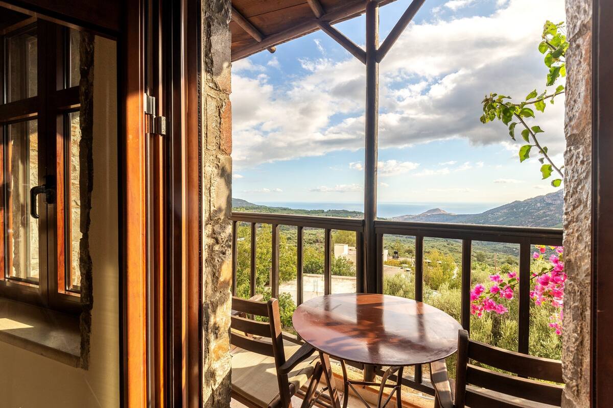 A balcony with a wooden table and chairs, overlooking a scenic landscape with mountains and vegetation.