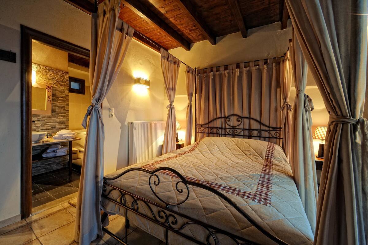 A four-poster bed with a quilt, a stone wall, a radiator, and a bathroom with a sink and towel rack.