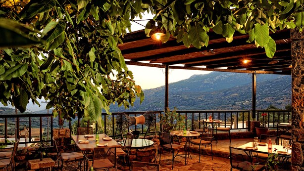 Un área de comedor al aire libre con mesas y sillas, rodeada de follaje y vistas a un paisaje montañoso.