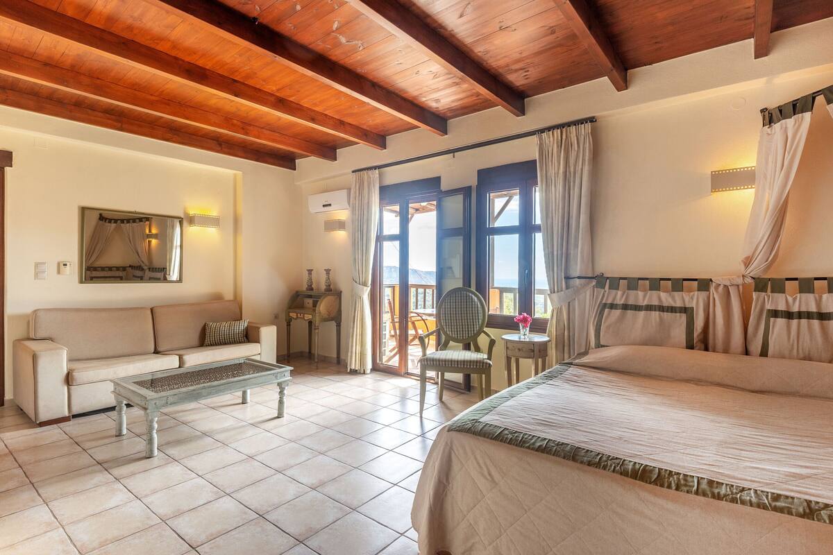 A bedroom with a bed, a sofa, a coffee table, and a chair. There is a window with curtains and a view of the outdoors.