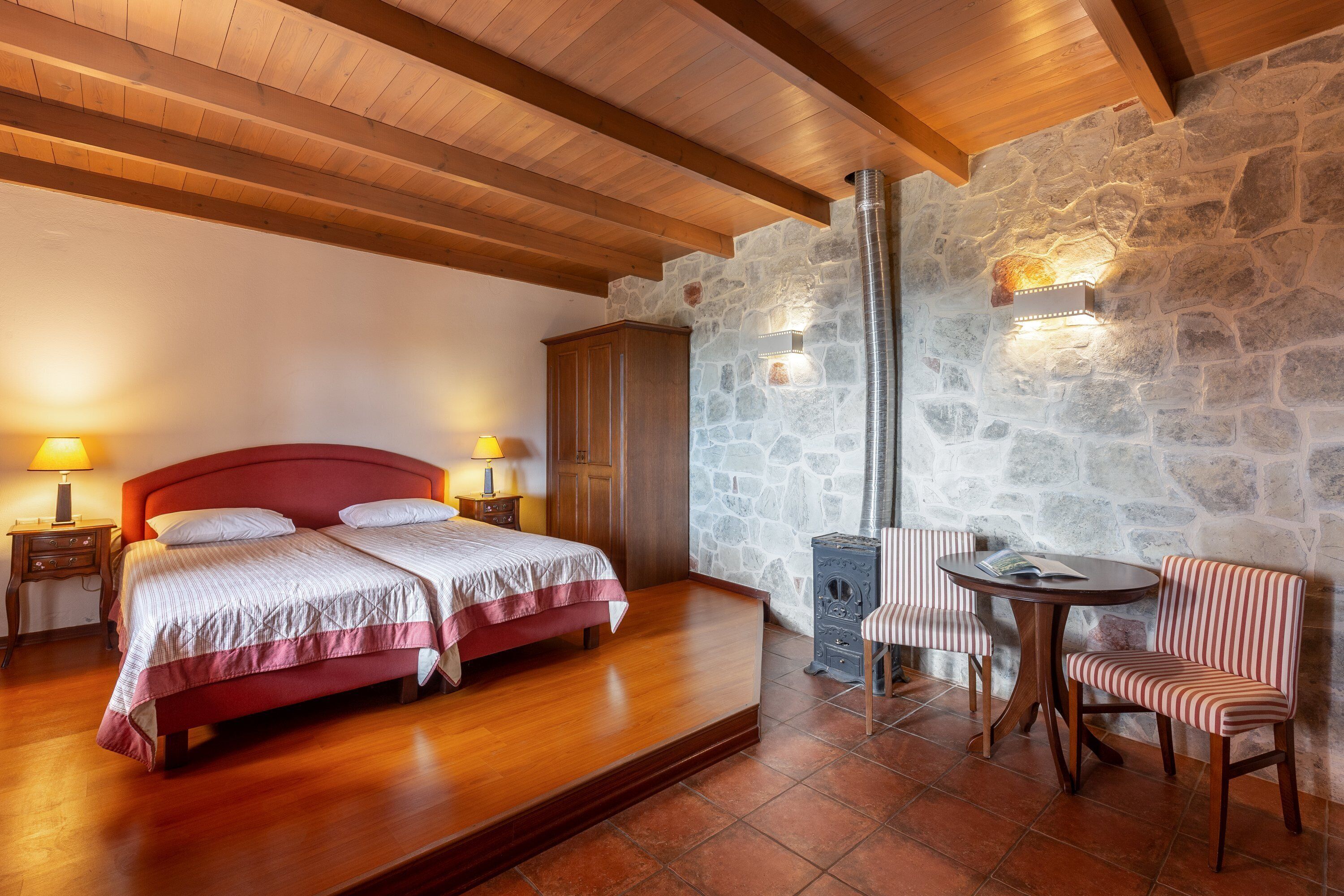 A bedroom with a wooden bed, a stone wall, a stone fireplace, a wooden table, and two chairs.