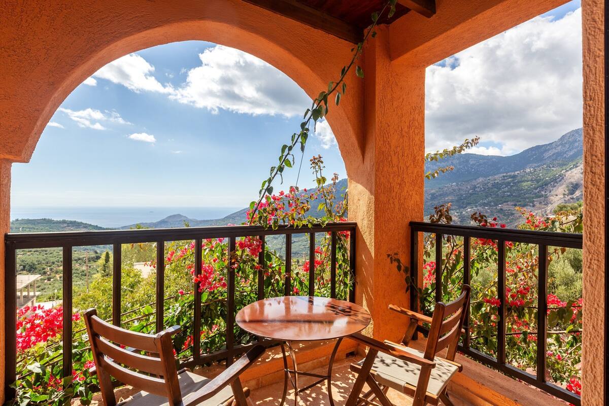 A balcony with a round table and chairs, surrounded by a black railing, overlooking a mountainous landscape with red flowers and greenery.