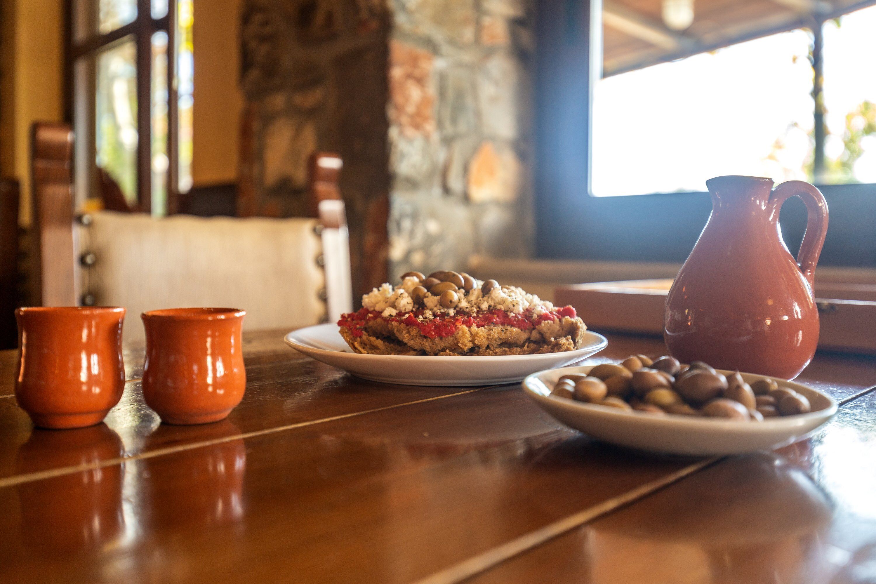 A wooden table with a plate of food, two small orange cups, and a red pitcher.