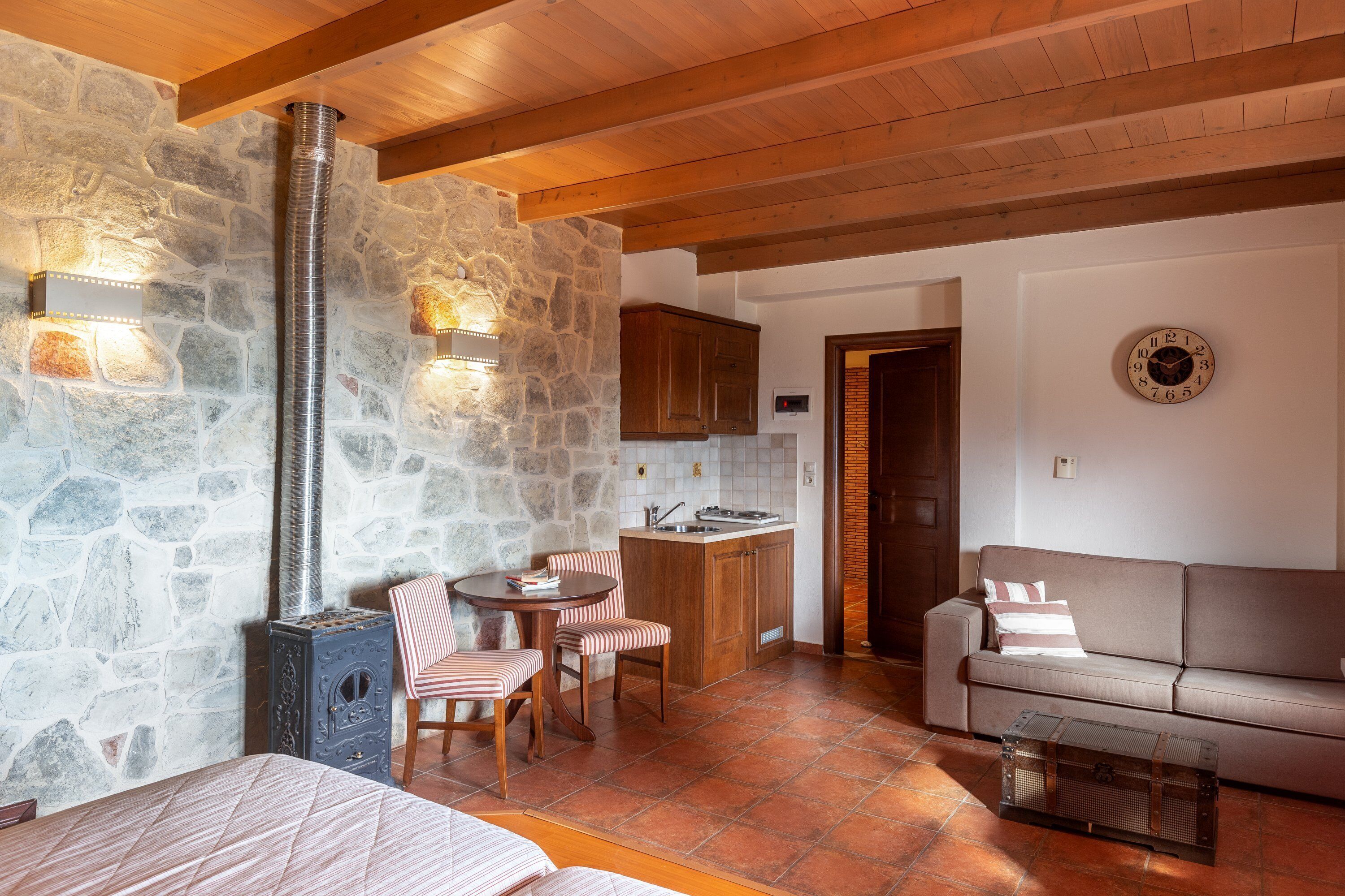Un intérieur chaleureux avec un mur en pierre, des poutres en bois au plafond, un coin repas avec des chaises à rayures, un canapé et un espace cuisine.