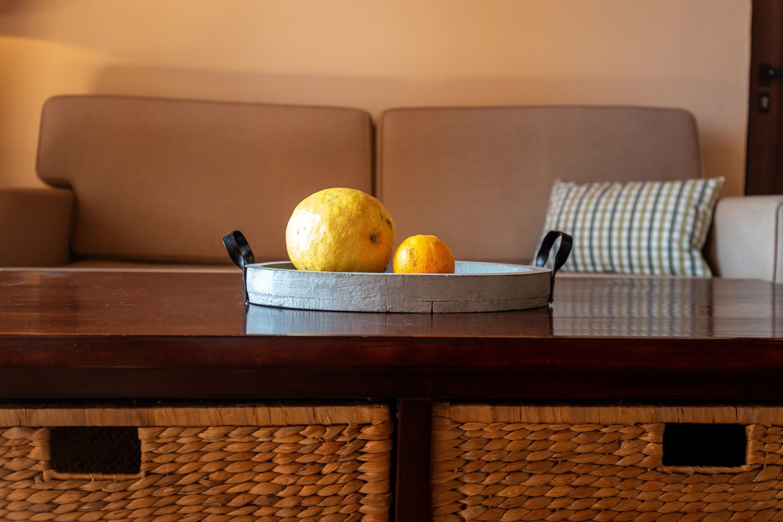 A round wooden tray with a lemon and an orange on a dark wooden table, placed on a brown sofa with a checkered pillow.