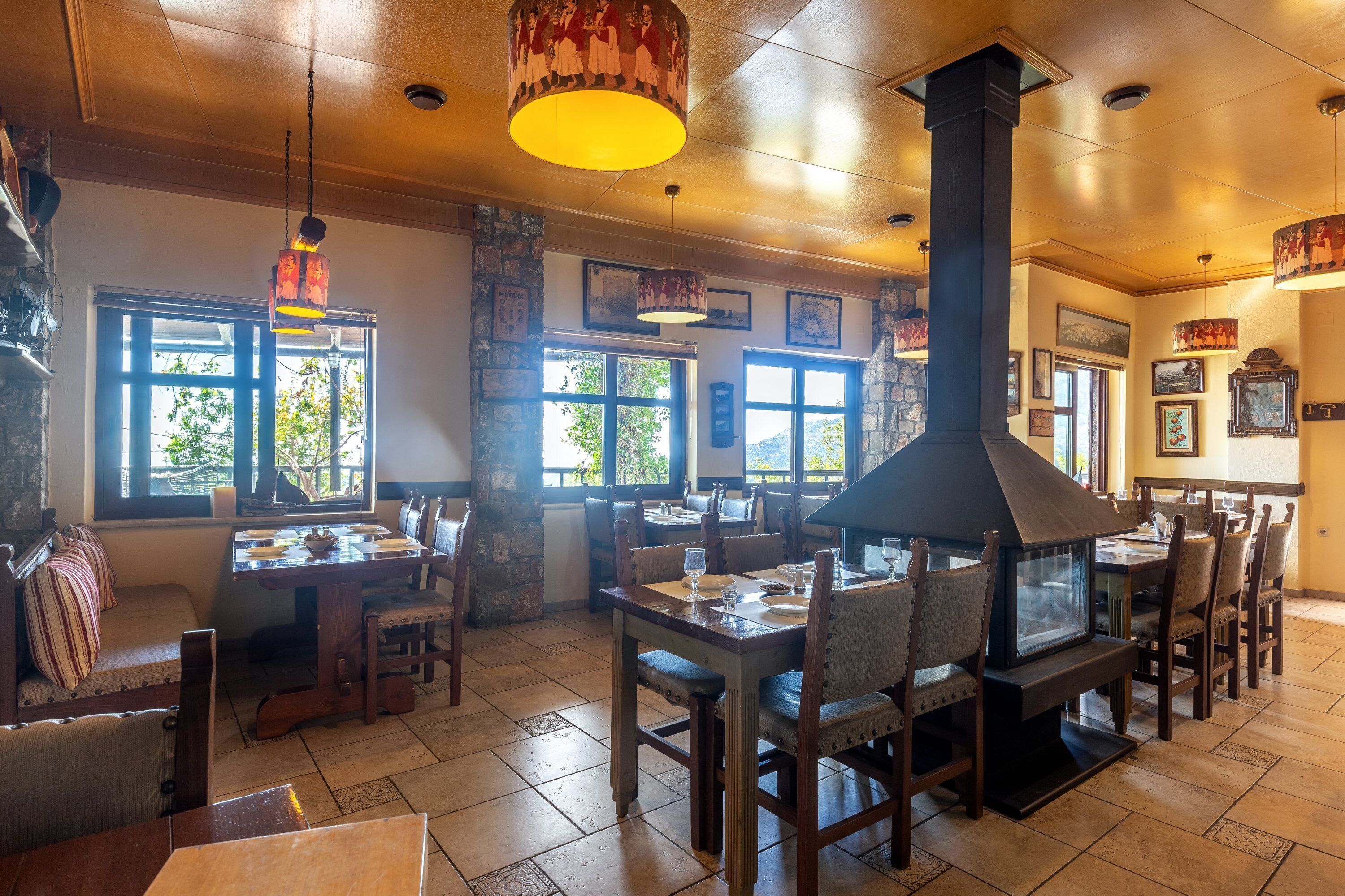 Un restaurante con mesas y sillas de madera, ventanas amplias y un horno de leña central.