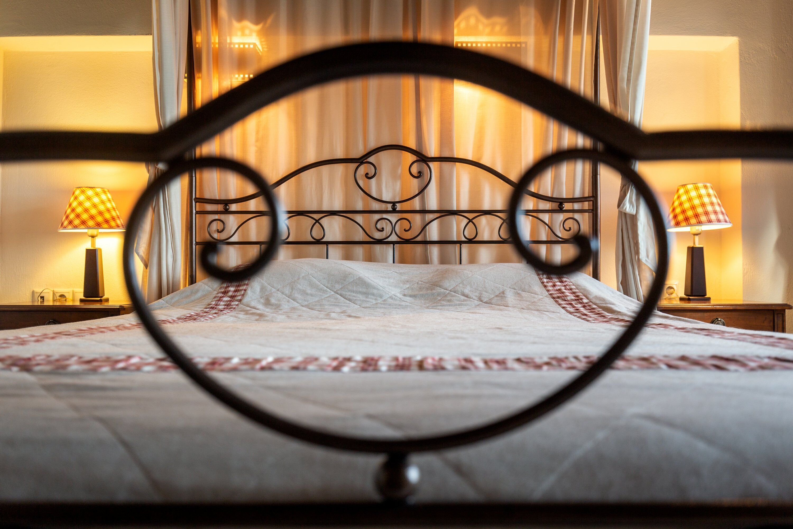 A bed with a metal headboard, two bedside lamps, and a view of a curtain and wall.