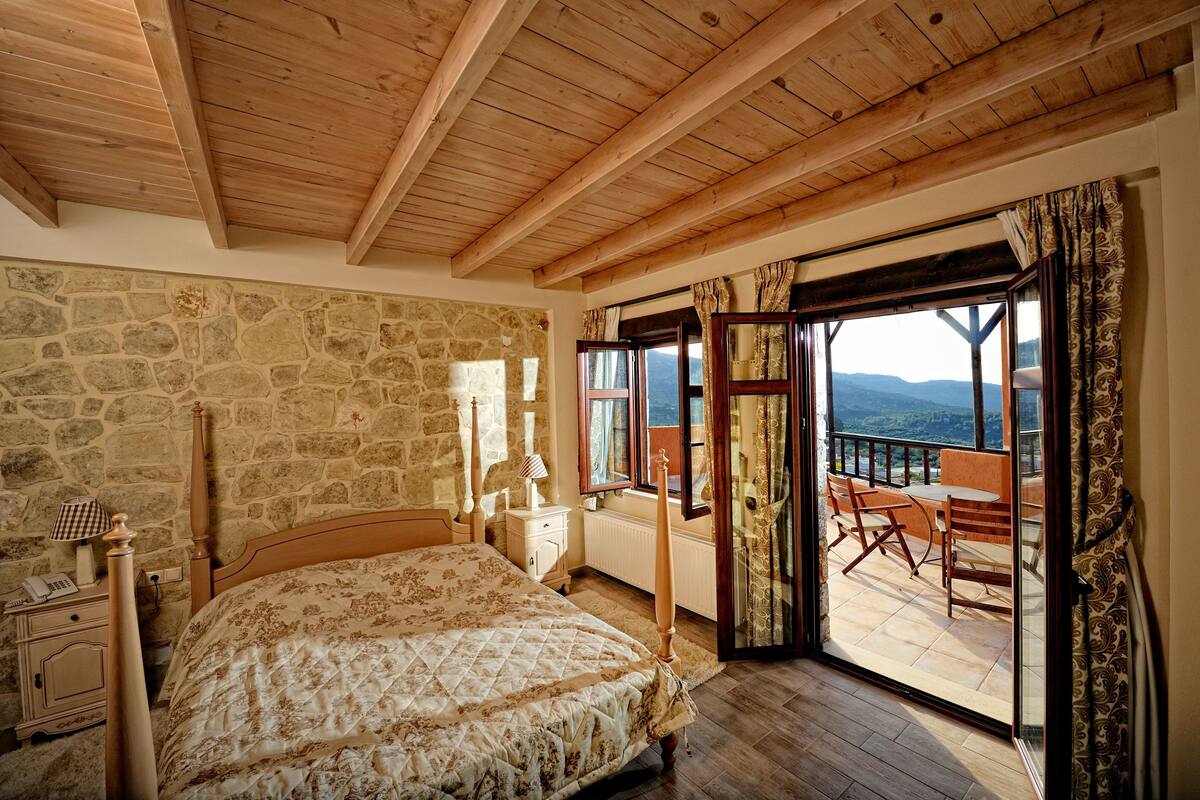 A bedroom with a stone wall, wooden ceiling beams, a bed with a wooden headboard, a nightstand with a lamp, and a balcony with a view of the mountains.