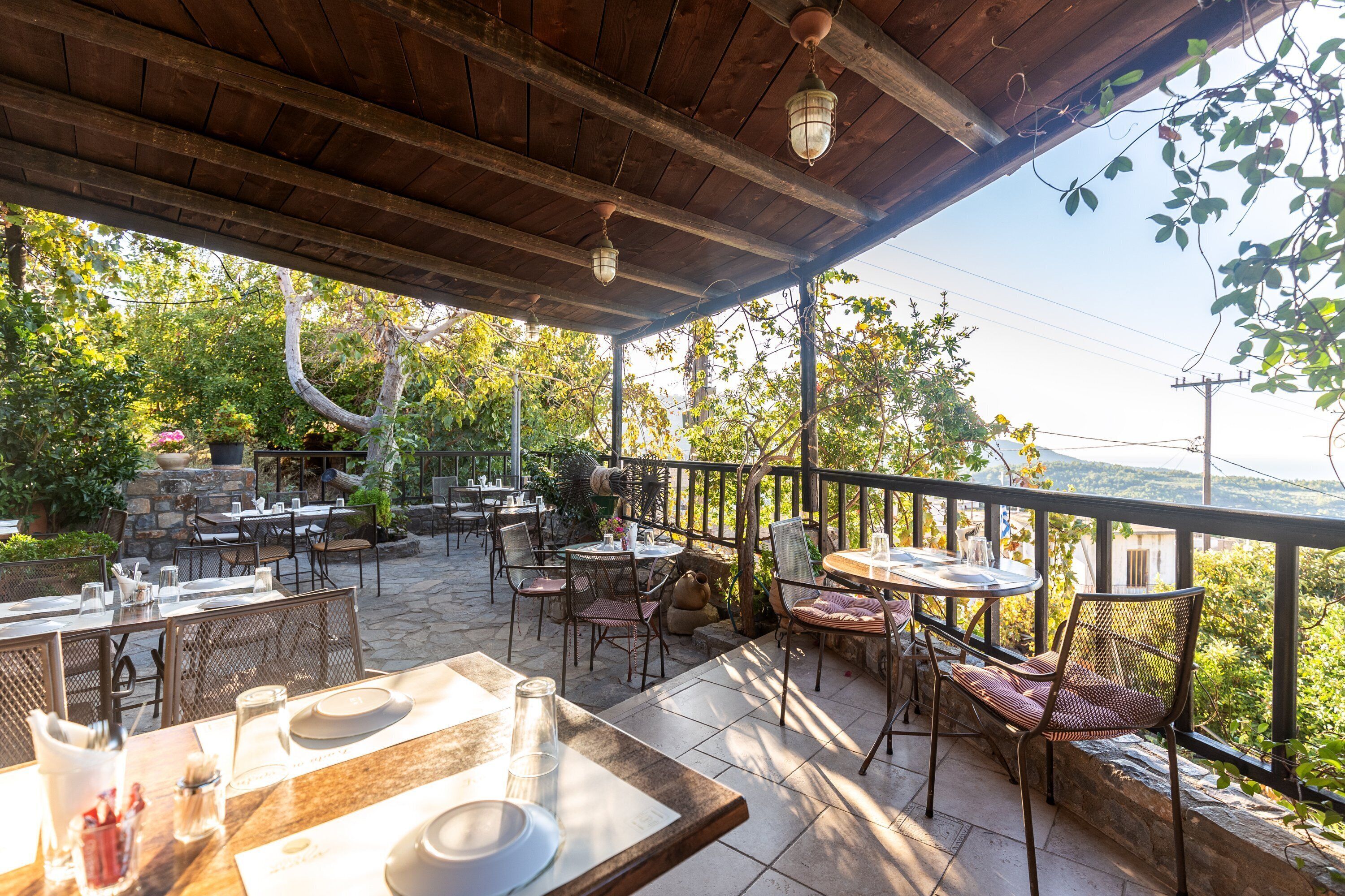 An outdoor patio with tables and chairs, a wooden ceiling, and a view of trees and hills.